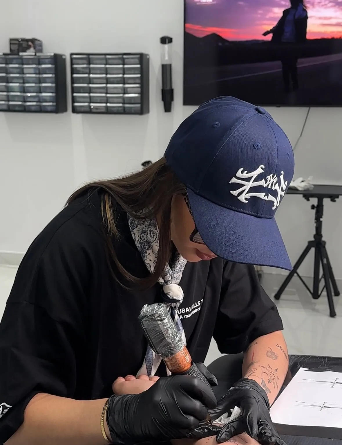 Gloved hands guiding a tattoo machine over a client's arm, a drawing of two swords on the table beside them.