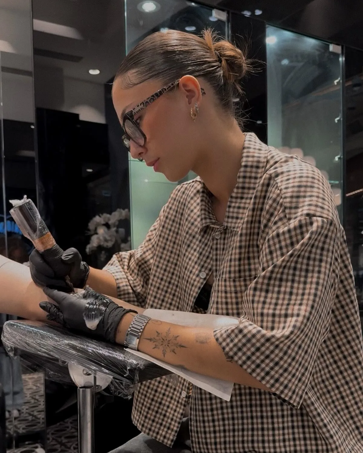 A tattoo artist mid-line, machine in hand, working on a client's arm in a glass-walled studio.